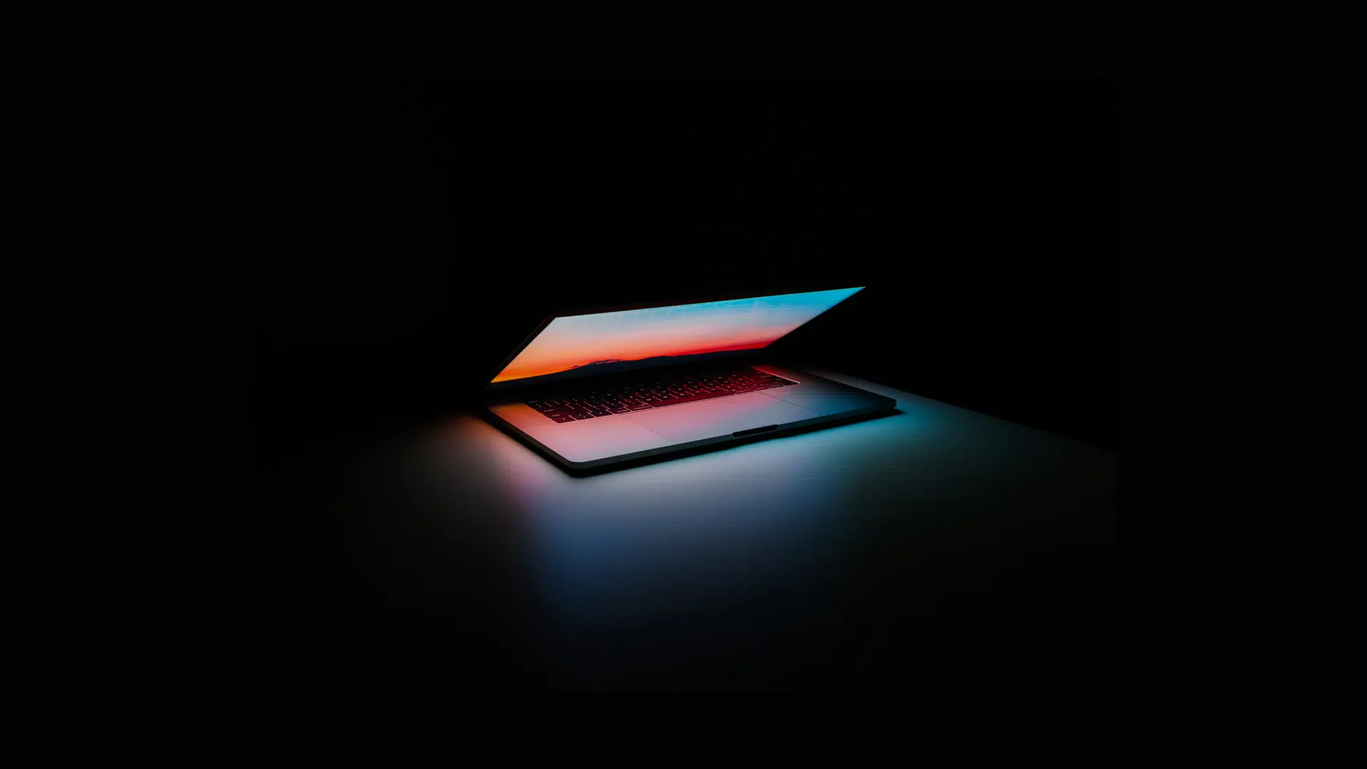 A partially open laptop on a dark surface emits a colorful glow from its screen, creating a contrast against the black background